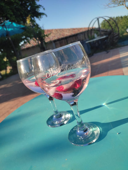 Two of our glasses on a blue table by our pool with our gin distillery barn in the background. There's raspberries in the glasses. French: Deux de nos verres sur une table bleue près de notre piscine avec notre grange-distillerie de gin en arrière-plan. Il y a des framboises dans les verres.