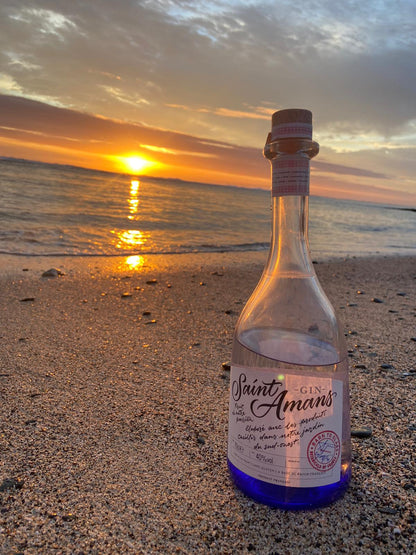 Our Saint Amans Original Gin, perfect drank pure, with tonic, or as a base for your cocktails, set against a beautiful southern French beach landscape at sunset. French: Notre Gin Original Saint Amans, parfait bu pur, avec du tonic ou comme base pour vos cocktails, sur  une belle plage de sable dans le sud de la France au coucher du soleil.