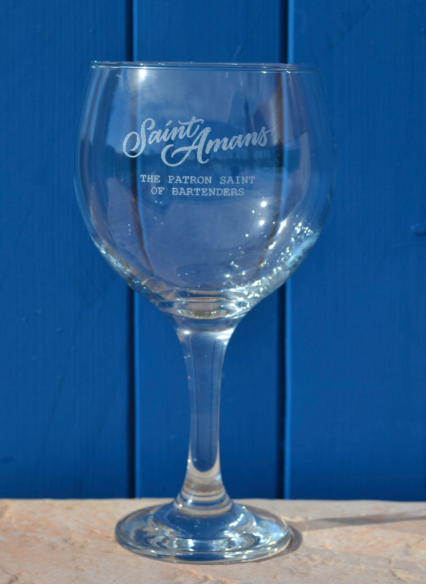 Our hand-engraved glass against a blue backdrop. The glass reads "The Patron Saint of Bartenders". French: Notre verre gravé à la main avec un arrière-plan bleu. On lit sur le verre « The Patron Saint of Bartenders ».